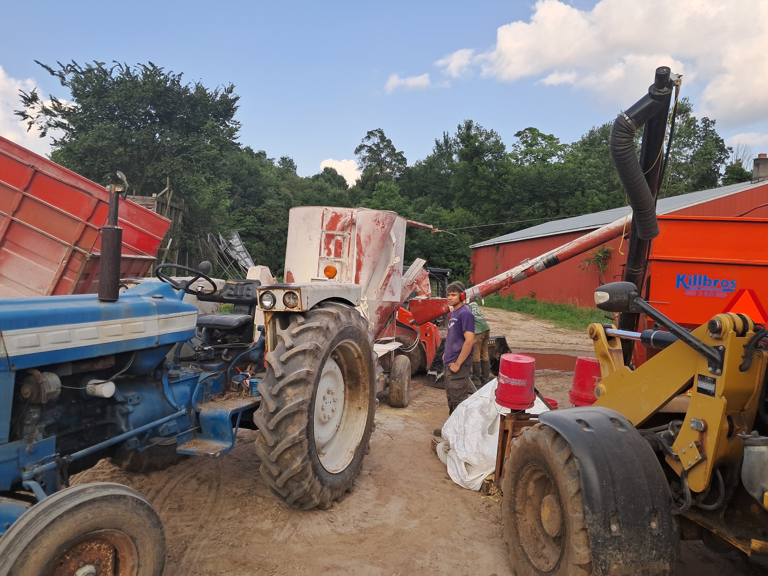 Farm equipment including blue and white tractors, a red trailer, and a yellow loader in a rural setting, with a man in a purple shirt standing nearby, surrounded by trees and a barn in the background.